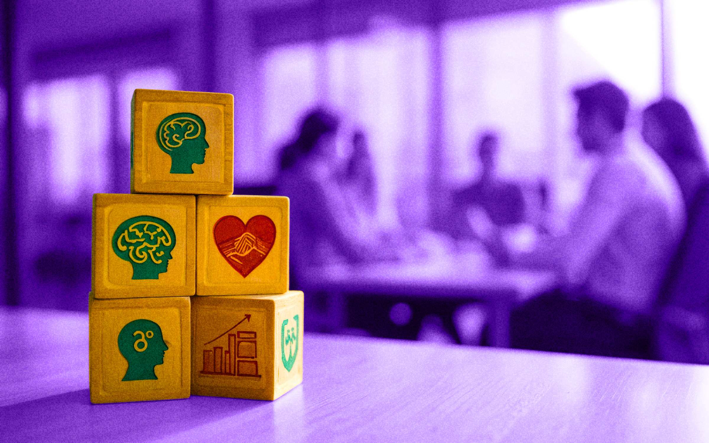 Stacked yellow blocks with icons of brains, a heart handshake, growth chart, and shield, set against people in discussion, symbolizing a mental health organization fostering collaboration, emotional intelligence, resilience, and protective support for wellbeing.